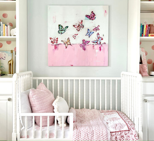 A canvas print by Chelsea McShane with a group of colorful butterflies of various sizes flying over a solid pink band at the bottom, with a white background above. The artwork is in a children's room above a toddler bed.